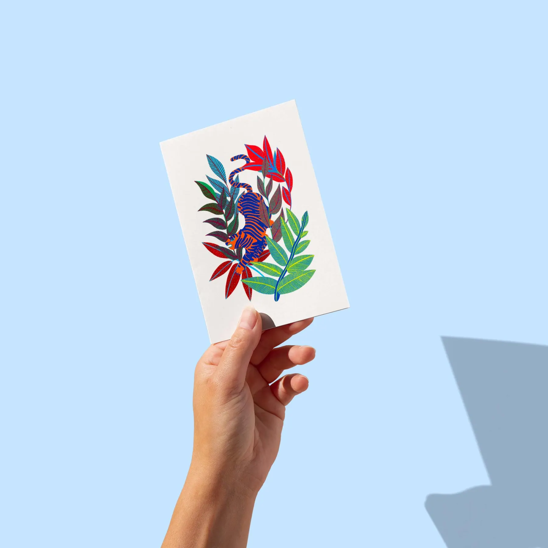 Tigre rayé bleu et orange lové dans un feuillage rouge et vert sur fond blanc, carte postale illustrée par Orane Sigal