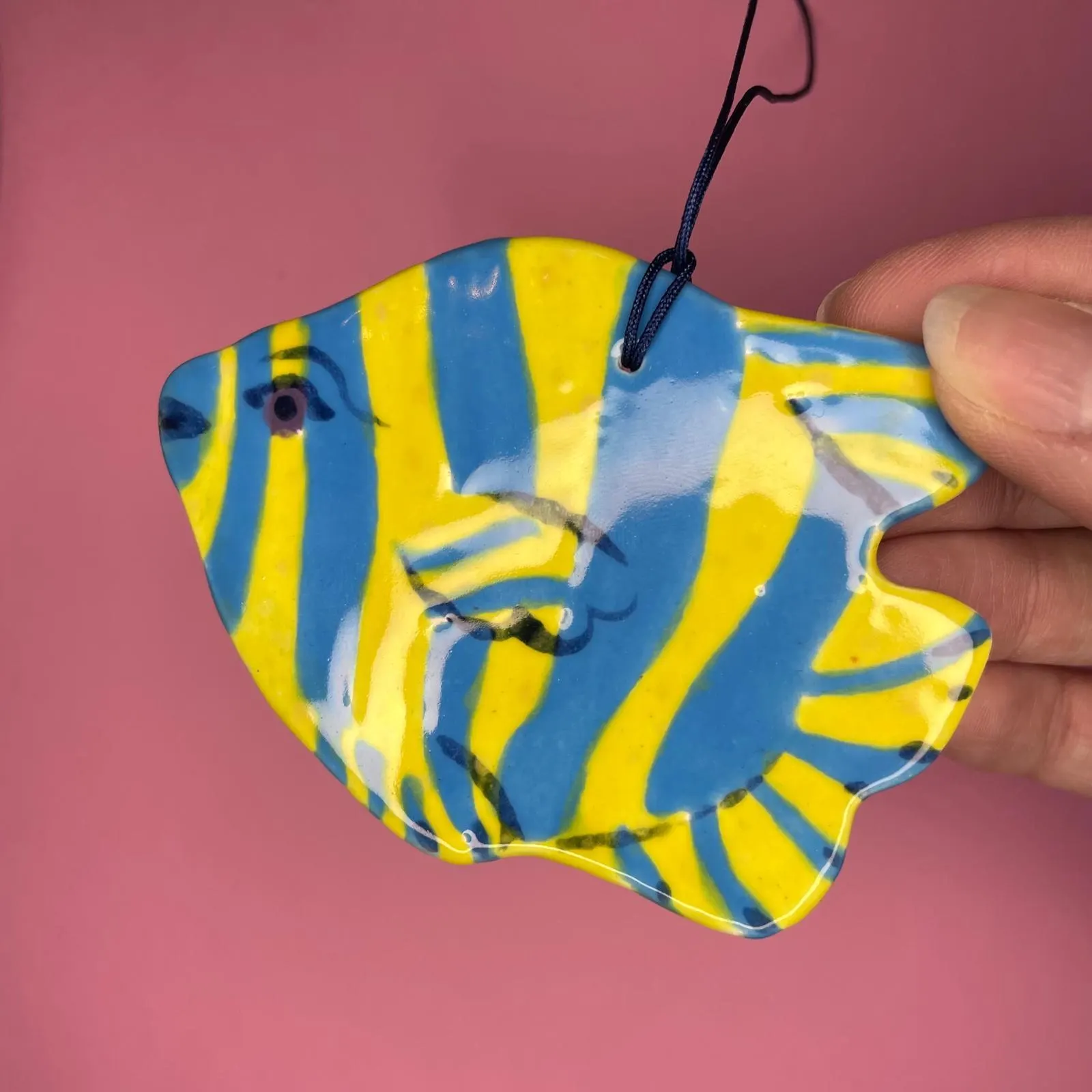 Suspension céramique en forme de poisson rayé jaune et bleu, fond rose, illustration par Orane Sigal